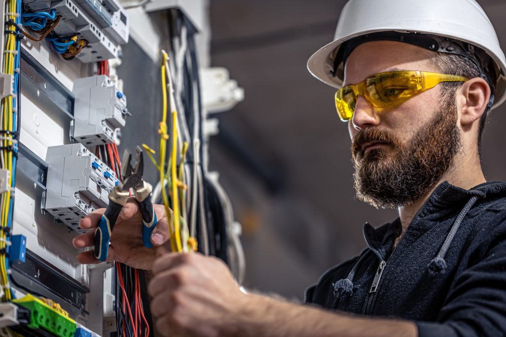 An electrician in a hard hat and safety glasses works on wiring in an electrical panel.